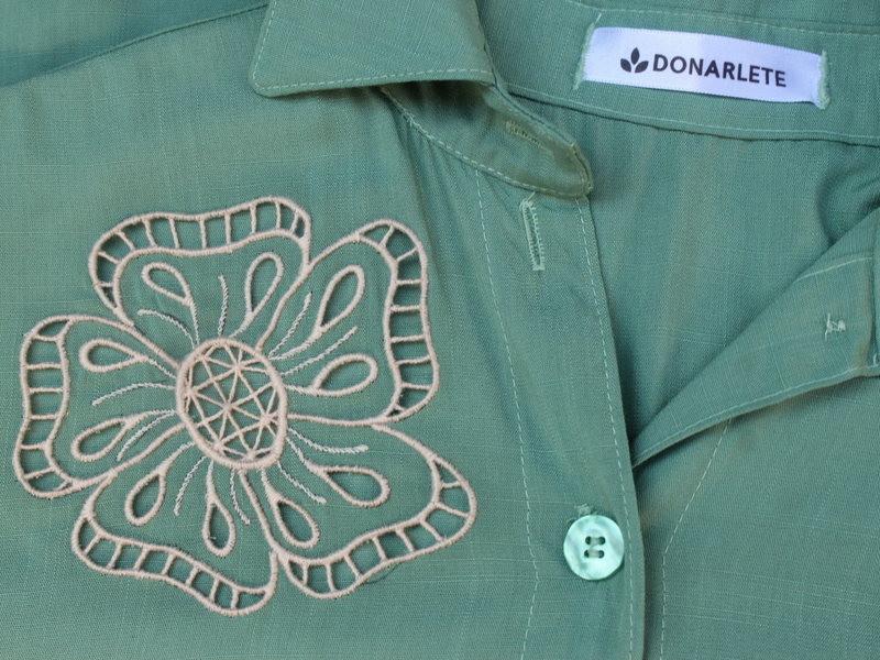 Detalhe de bordado em ponto de flor sobre camisa verde de alfaiataria, com etiqueta da marca Donarlete